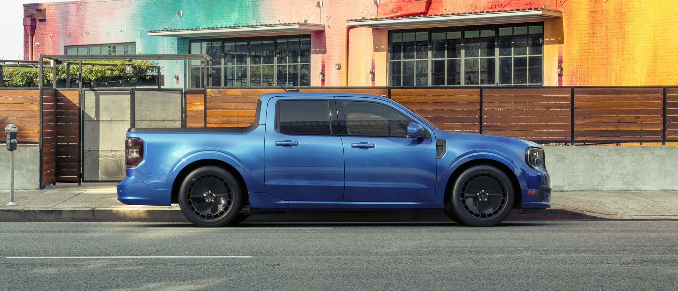 A side profile of a blue Ford Maverick parked on a city street against a vibrant building backdrop, emphasizing its sleek and modern design for urban driving.